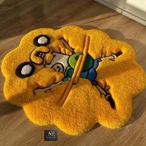 Pode incluir: Um tapete tufado amarelo em forma de nuvem com um gráfico da personagem de desenho animado Jake o Cão, com Finn o Humano parcialmente visível em roupas azuis e verdes. Um pequeno logótipo quadrado preto com o texto NUMAIR ENTERPRISES está presente.