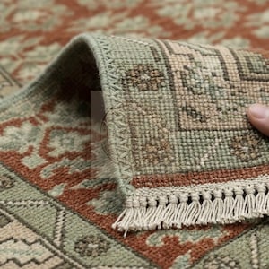 May include: A woven area rug featuring a repeating floral and geometric pattern in shades of sage green, terracotta orange, and cream. The rug has a finished edge with a decorative stitched border and a short fringe along the bottom edge.