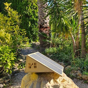 May include: A wooden slant board with a black textured surface, resting on a rock in a garden setting. The board has the text "SLANT BOARD" printed on it. The background features lush greenery and palm trees.