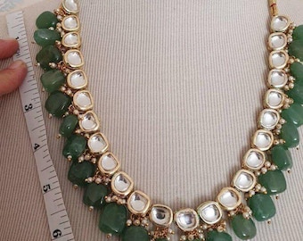Puede incluir: Un collar dorado con cuentas de piedras preciosas verdes y detalles cuadrados blancos. El collar se exhibe en un maniquí. El collar mide aproximadamente 15 cm de largo.