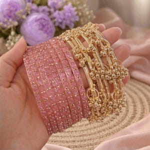 Könnte beinhalten: Eine Hand präsentiert eine Sammlung goldener und rosa Armreifen. Die rosa Armreifen sind einfarbig mit kleinen, dekorativen Akzenten. Die goldenen Armreifen haben ein Design mit hängenden Glöckchen. Der Hintergrund umfasst lila Blumen und einen rosa Stoff.