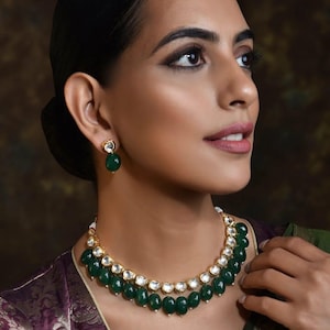 May include: A woman wearing a gold necklace and matching earrings. The necklace has two strands of dark green beads with clear, round stones. The earrings have a similar design, with a single green bead. The woman is wearing a purple and green garment.