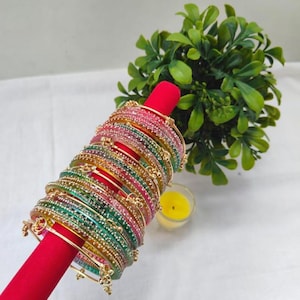 May include: A collection of colorful bangles, displayed on a red stand. The bangles are adorned with small, sparkling embellishments and come in shades of pink, green, yellow, and gold. A small green plant and a yellow candle are in the background.