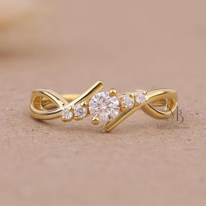 Op de afbeelding: Een gouden ring met een centrale ronde briljant geslepen heldere steen, geflankeerd door twee kleinere heldere stenen aan elke kant. De band heeft een gekruist ontwerp van glad goud. De tekst MettleBloom is zichtbaar aan de rechterkant.