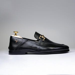 May include: Black leather loafer with a gold-colored buckle and a small emblem. The shoe has a textured surface and a classic design. A wooden shoe tree with a gold-colored top is inside the shoe.