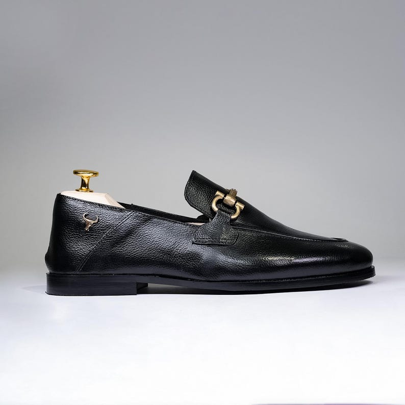 May include: Black leather loafer with a gold-colored buckle and a small emblem. The shoe has a textured surface and a classic design. A wooden shoe tree with a gold-colored top is inside the shoe.