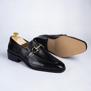 May include: Black leather loafers with a gold-colored buckle detail. One shoe is upright, and the other is lying on its side, revealing the tan sole. The shoes have a low heel and a classic design.