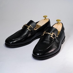 May include: A pair of black leather loafers with gold-colored hardware. The shoes have a classic design with a rounded toe and a decorative buckle. Shoe trees are inserted inside the shoes to maintain their shape.