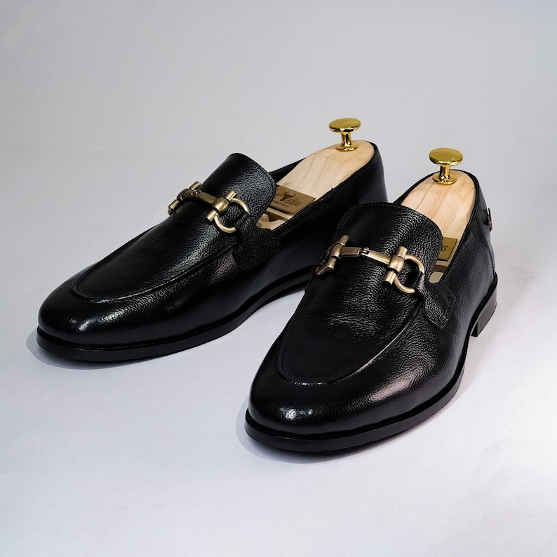 May include: A pair of black leather loafers with gold-colored hardware. The shoes have a classic design with a rounded toe and a decorative buckle. Shoe trees are inserted inside the shoes to maintain their shape.