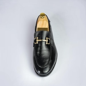 May include: Black leather loafer with a gold-tone buckle. The shoe has a rounded toe and a textured leather upper. The shoe is on a wooden shoe tree. The overall style is classic and elegant.