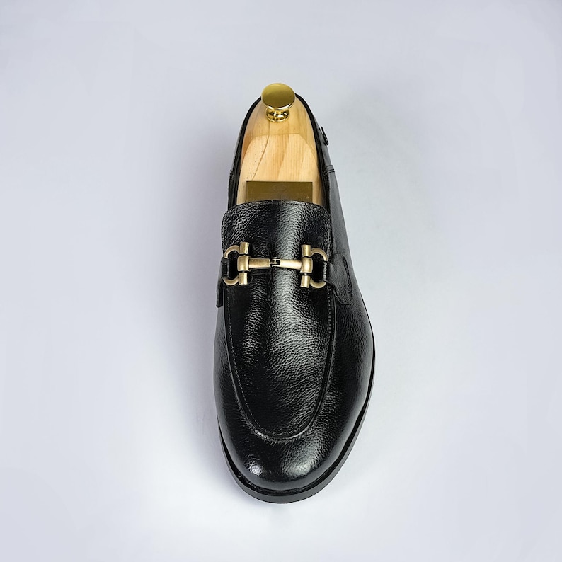 May include: Black leather loafer with a gold-tone buckle. The shoe has a rounded toe and a textured leather upper. The shoe is on a wooden shoe tree. The overall style is classic and elegant.