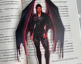 Puede incluir: Figura acrílica de un personaje masculino con grandes alas detalladas. Viste un traje negro con detalles rojos. La figura está colocada frente a un libro abierto, con una base reflectante. Las alas son intrincadas.
