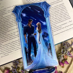 Peut inclure: Un présentoir de livre en acrylique bleu et blanc avec une illustration romantique d'un couple sous un ciel étoilé. Le présentoir est devant un livre ouvert avec du texte.