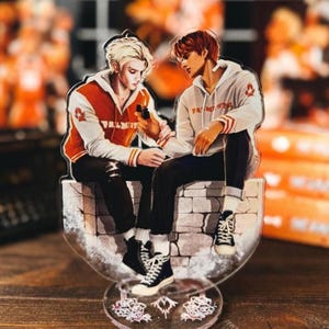 May include: A clear acrylic figure featuring two anime-style characters sitting on a stone wall. One character wears a red and white jacket, the other a white hoodie. Both wear black pants and sneakers. The base has a floral design.