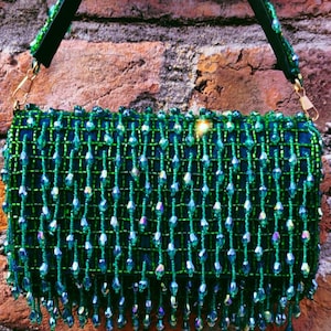 Puede incluir: Un bolso de mano verde con cuentas con una correa negra y herrajes dorados. El bolso es rectangular con un fleco de cuentas colgantes. El fondo es una pared de ladrillos.