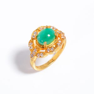 Puede incluir: Anillo dorado con una piedra verde ovalada en el centro. La piedra está rodeada por un halo de piedras más pequeñas y claras, dispuestas en un diseño floral. La banda del anillo también es dorada, creando una apariencia lujosa.