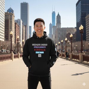 Puede incluir: Una sudadera negra con las palabras "Comparte Bondad, Spread Kindness, 把善意傳出去, Szerz Dobro" en un degradado de colores. La sudadera tiene un bolsillo delantero y un código QR. El fondo muestra una ciudad con edificios altos.