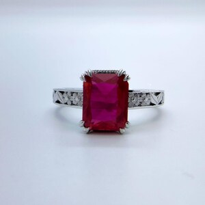 May include: A silver ring with a large, rectangular, ruby-red gemstone. The band features a floral pattern. The gemstone is set in a claw-like setting. The ring is displayed on a white surface.