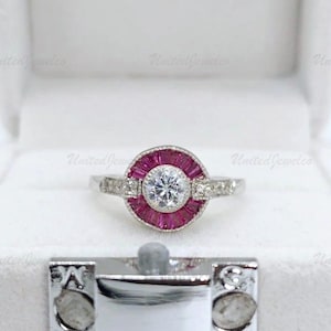May include: A silver ring with a central round, clear gemstone surrounded by a halo of small, rectangular, red gemstones. The ring's band features small, clear gemstones on either side of the central design. The ring is displayed in a white jewelry box.