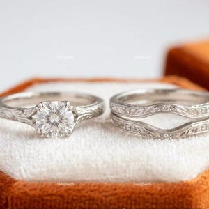Peut inclure: Une bague de fiançailles argentée avec une pierre centrale ronde taille brillant et une alliance assortie profilée. Les deux anneaux présentent des motifs de feuilles gravés complexes, l'alliance étant ornée de petites pierres d'accent.