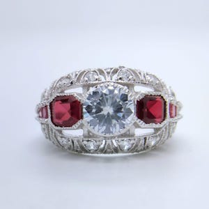 May include: A silver ring with a large, round, clear central stone, framed by two square, deep red gems. The ring showcases detailed filigree work and small, clear accent stones, offering a vintage aesthetic.