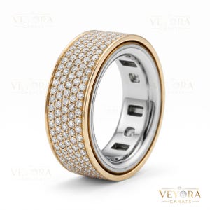 May include: A wide, two-tone ring with a gold band and a silver interior. The outer band is encrusted with numerous small, sparkling stones. The ring is set against a white background, highlighting its design and craftsmanship.