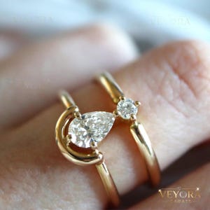 May include: Two gold rings with diamonds. One ring features a pear-shaped diamond, and the other has a round diamond. The rings are set in gold and are on a finger.