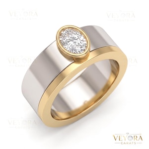 May include: A two-tone ring featuring a wide silver-coloured band with a gold-coloured edge and a central oval-shaped bezel setting. The setting holds a single, clear, faceted oval gemstone. The brand name VEYORA CARATS is visible in the corner.