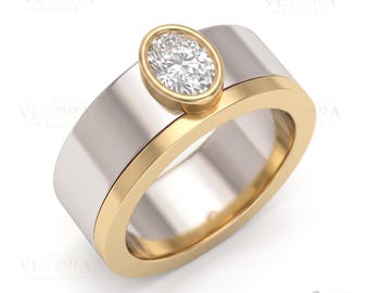 May include: A two-tone ring featuring a wide silver-colored band with a gold-colored edge and a central oval-shaped bezel setting. The setting holds a single, clear, faceted oval gemstone. The brand name VEYORA CARATS is visible in the corner.