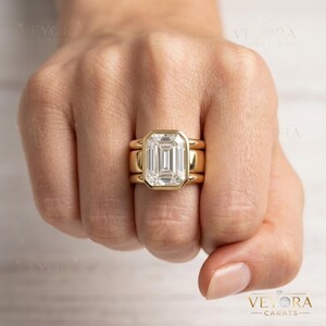 May include: A gold ring featuring a large, rectangular emerald-cut center stone set in a bezel setting. The ring is composed of a thick, smooth gold band that appears to be a stack of three connected rings, creating a wide, bold profile on the finger.