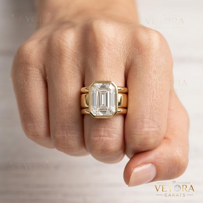 May include: A gold ring featuring a large, rectangular emerald-cut center stone set in a bezel setting. The ring is composed of a thick, smooth gold band that appears to be a stack of three connected rings, creating a wide, bold profile on the finger.