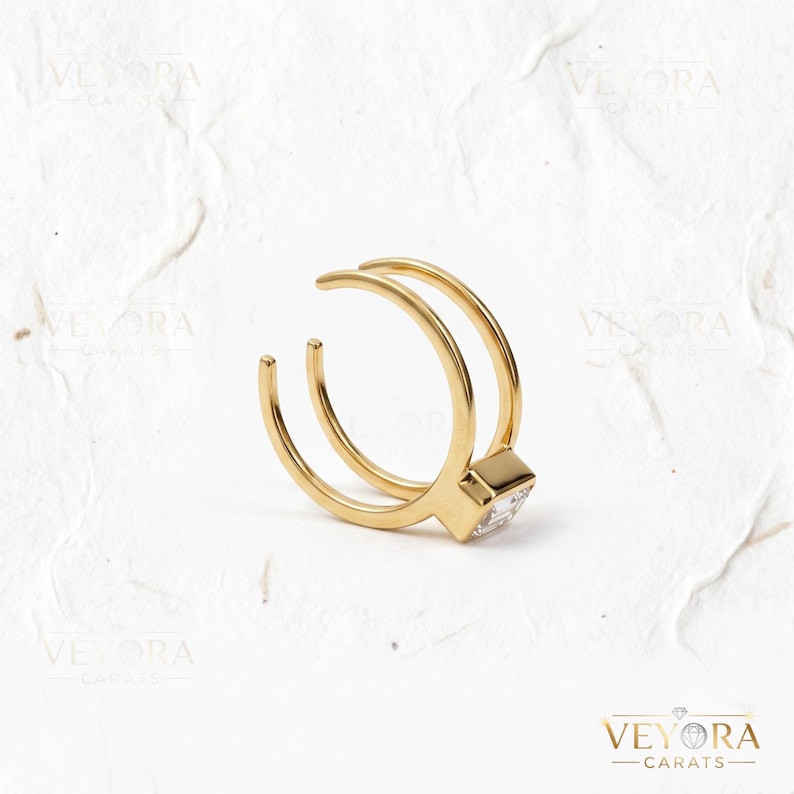 May include: A gold-toned open ring featuring a double band design that converges at a single point. A clear, rectangular-cut stone is set into the metal at the point where the two bands meet. The ring has a polished finish and a minimalist structure.
