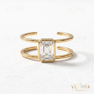 May include: A gold open-band ring featuring a central emerald-cut clear gemstone held in a bezel setting. The band consists of two parallel gold wires that connect to the rectangular stone, creating a split-shank design with a polished finish.