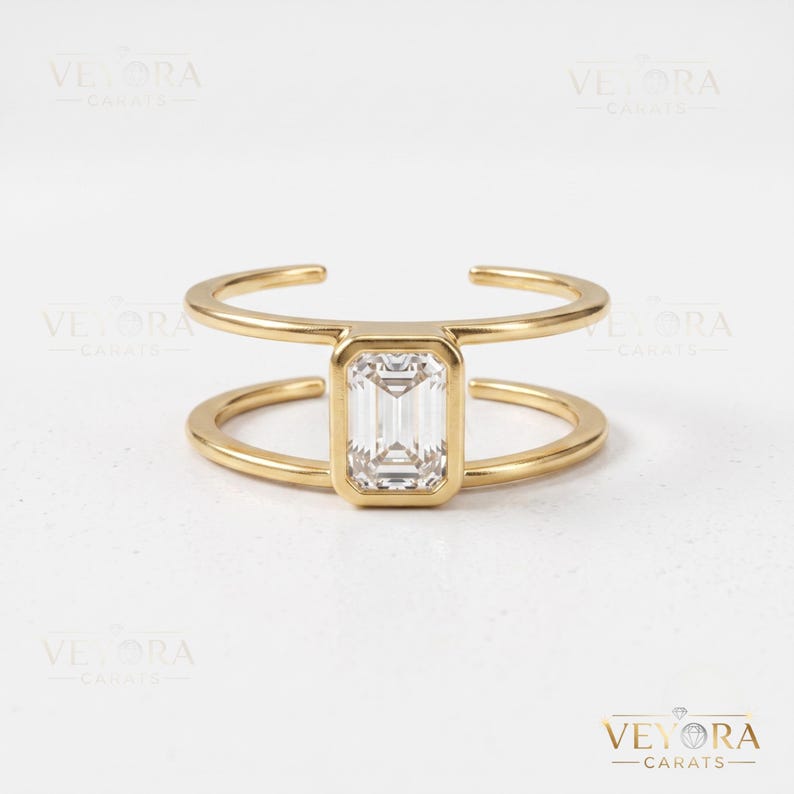 May include: A gold-toned open ring featuring a central emerald-cut clear stone set in a rectangular bezel. The band consists of two parallel gold-toned wires that connect at the stone, creating a split-shank design with a polished metallic finish.