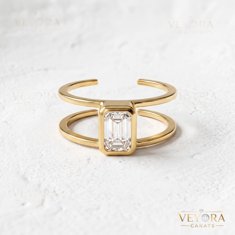 May include: A gold ring featuring a double band design and a central emerald-cut clear gemstone set in a bezel setting. The ring has an open-ended structure and a polished finish. The text VEYORA CARATS is visible in the bottom right corner.