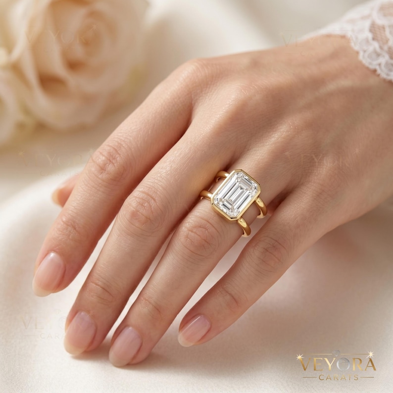May include: A gold ring featuring a large emerald-cut clear gemstone set in a bezel setting. The band is split into two distinct gold bars that connect to the central stone setting, creating an open, architectural design on the finger.
