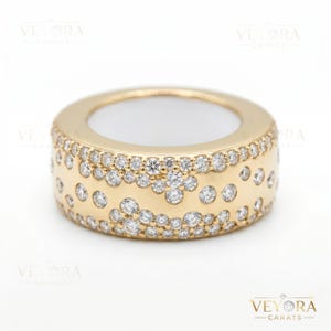 Puede incluir: Un anillo ancho, de color dorado, con una banda de pequeños diamantes brillantes. El anillo tiene una banda gruesa con una serie de diamantes engastados en un patrón alrededor de la superficie exterior. El interior del anillo es blanco. El anillo es de Veyora Carats.