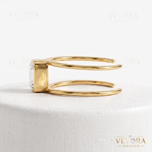 May include: A gold ring featuring a rectangular emerald-cut clear stone set in a bezel setting. The band is open and consists of two parallel gold wires that connect to the stone setting on one side, creating a modern and geometric appearance.