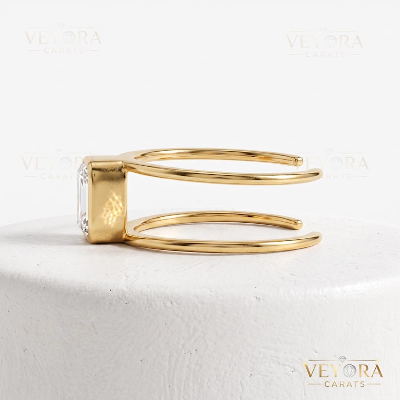 May include: A gold ring featuring a rectangular emerald-cut clear stone set in a bezel setting. The band is open and consists of two parallel gold wires that connect to the stone setting on one side, creating a modern and geometric appearance.