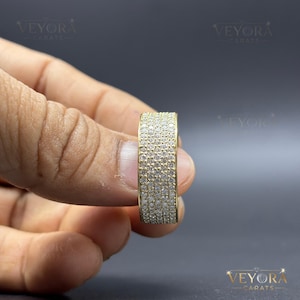 Könnte beinhalten: Ein breiter Goldring mit Pavé-Fassung, bei dem mehrere Reihen kleiner, klarer, rund geschliffener Steine die gesamte Außenfläche bedecken. Der Ring ist so positioniert, dass die detaillierte Textur der Steine und das Goldfinish sichtbar sind.