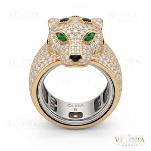 May include: A gold ring featuring a panther head design, encrusted with numerous small diamonds. The panther's eyes are emerald green, and its nose is black. The ring has a silver interior with the text "OURA 5".