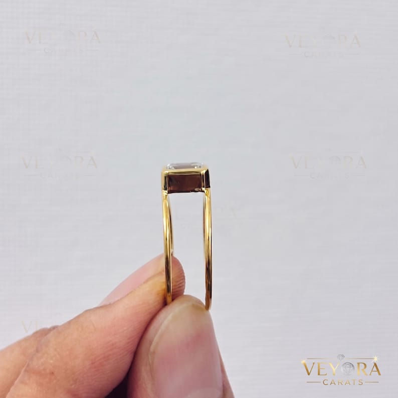 May include: A gold ring featuring a rectangular, clear gemstone set in a high-profile bezel setting. The band is thin and smooth, showcasing a side profile view of the stone's depth and the metal construction of the setting.
