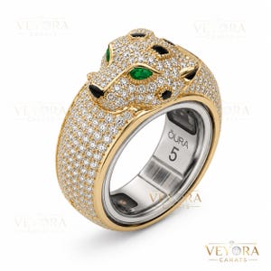 Puede incluir: Un anillo de oro con un diseño de cabeza de pantera, incrustado con pequeños diamantes brillantes. Los ojos de la pantera son de color verde esmeralda y tiene manchas negras. El anillo tiene el texto "OURA" y el número "5" grabado en el interior.