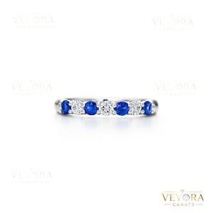 May include: A silver ring with alternating round, brilliant-cut blue sapphires and white diamonds. The gemstones are set in a row across the top of the ring. The ring is displayed on a white background.