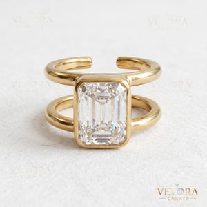 May include: A gold-toned open-band ring featuring a large, rectangular emerald-cut clear stone set in the center. The band consists of two parallel gold-toned metal bars that connect to the stone setting, creating a modern, open-style design.