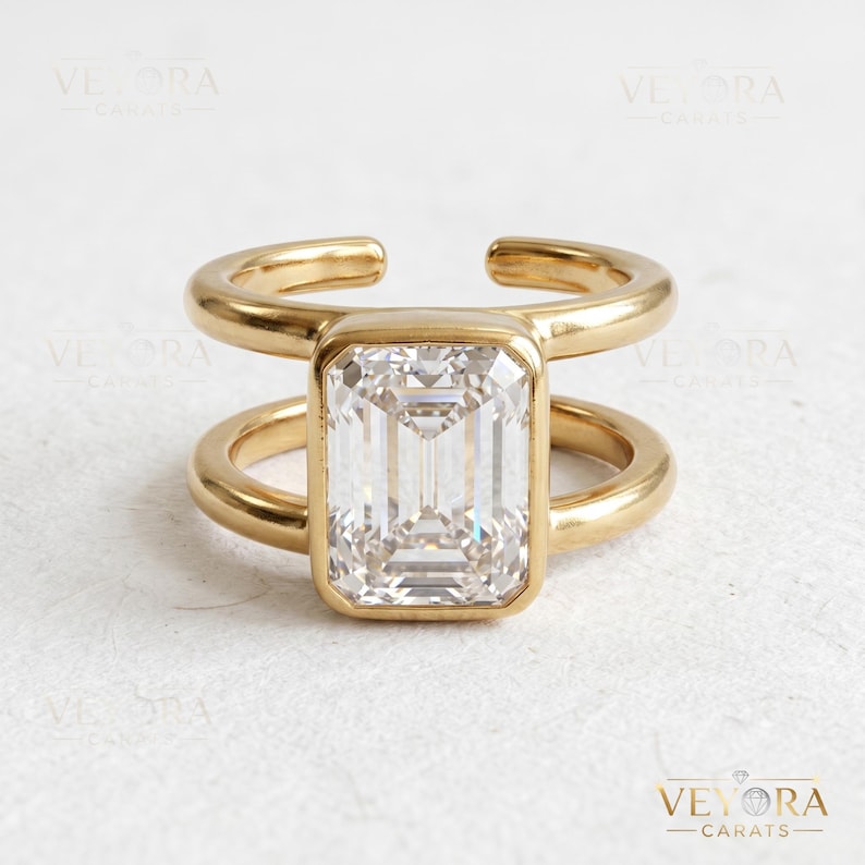 May include: A gold-toned open-band ring featuring a large, rectangular emerald-cut clear stone set in the center. The band consists of two parallel gold-toned metal bars that connect to the stone setting, creating a modern, open-style design.