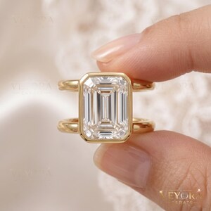 May include: A gold ring featuring a large, rectangular emerald-cut clear gemstone set in a bezel setting. The band is split into two parallel gold bars that connect to the central stone setting, creating a modern, open-frame design.