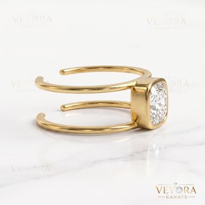 May include: A gold ring featuring a cushion-cut clear gemstone set in a bezel setting. The band is open and consists of two parallel gold wires that connect to the central stone setting, creating a split-shank design with a polished finish.