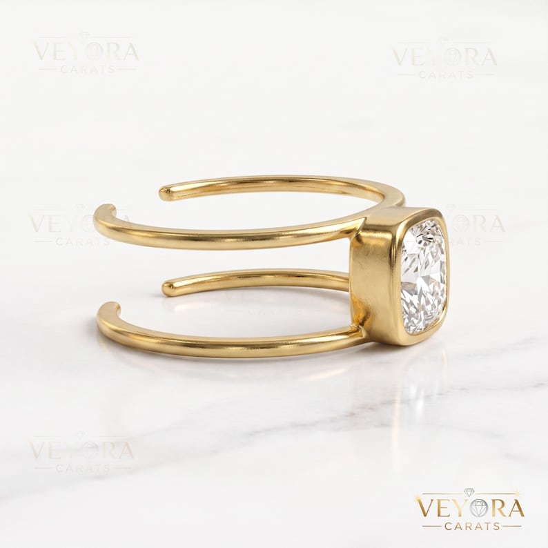 May include: A gold ring featuring a cushion-cut clear gemstone set in a bezel setting. The band is open and consists of two parallel gold wires that connect to the central stone setting, creating a split-shank design with a polished finish.