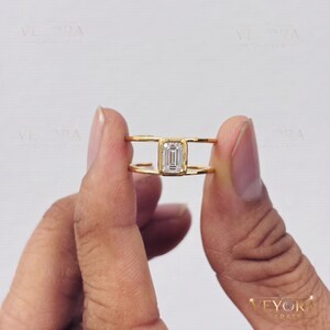 May include: A gold ring featuring a central emerald-cut clear gemstone held in a bezel setting. The band consists of two parallel gold wires that connect at the sides to the central stone setting, creating an open, split-shank design.